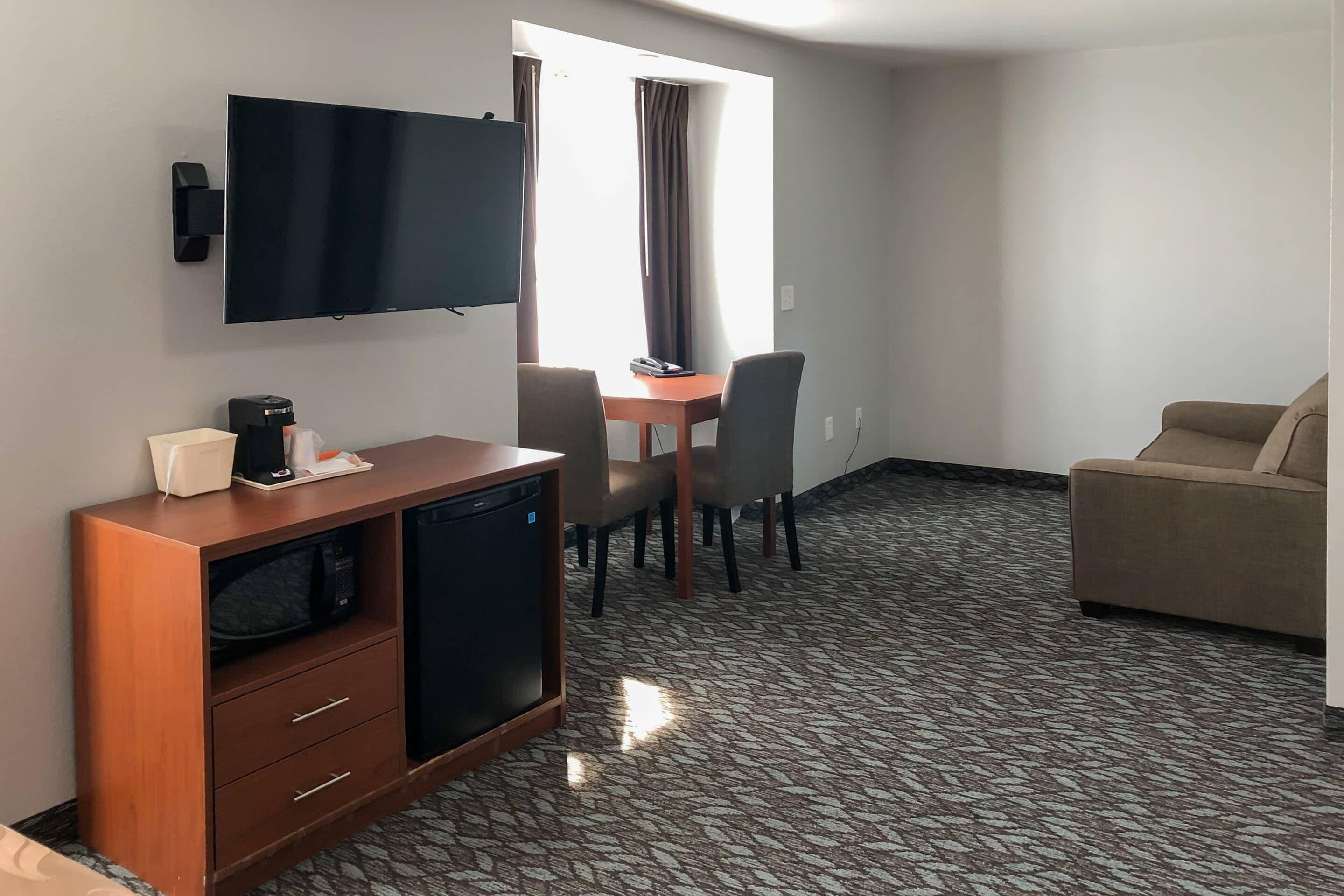 A hotel room with a flat-screen TV mounted on the wall, a small wooden desk with a microwave, a coffee maker, and a chair. There is a sofa and a window with curtains.