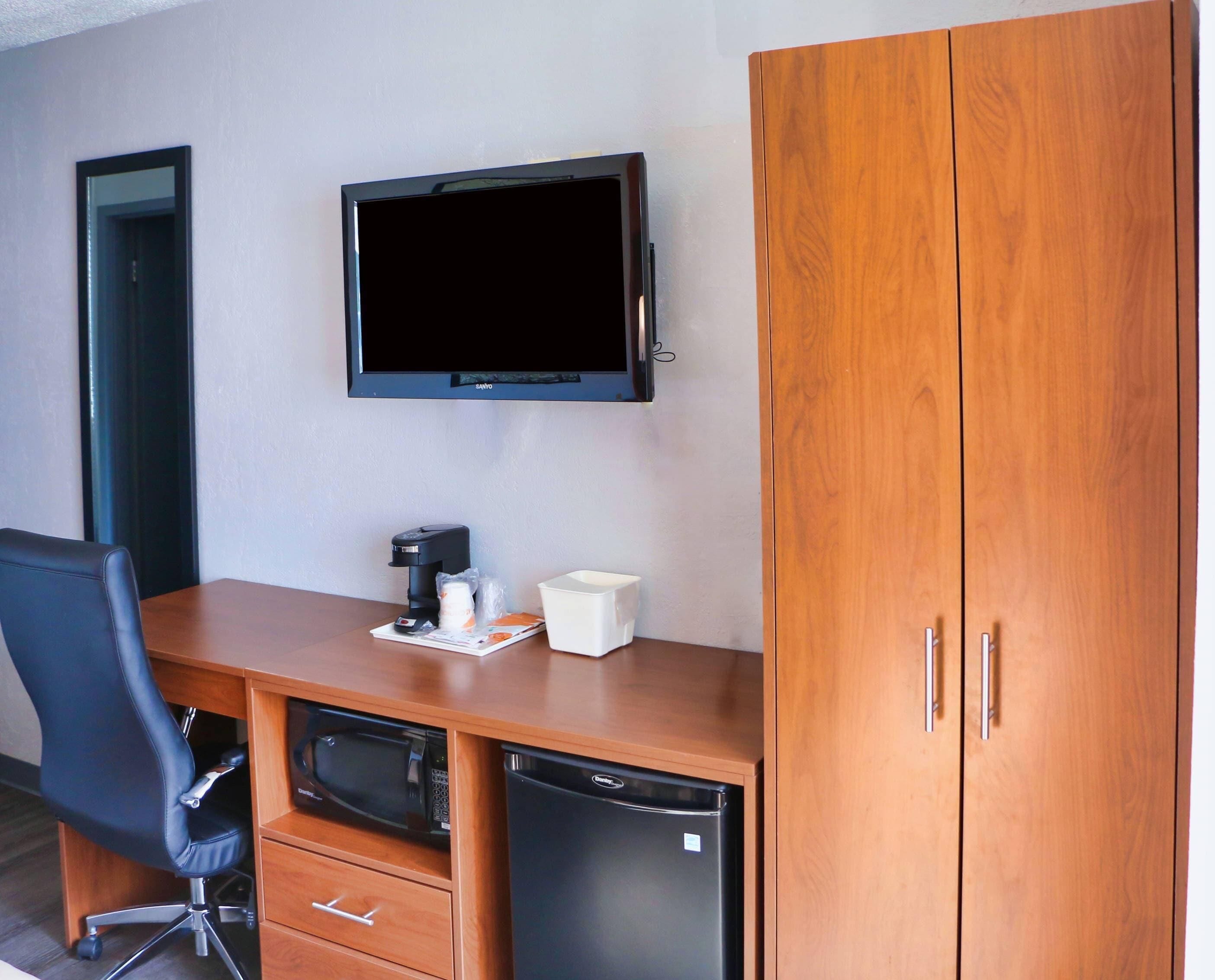 Habitación de hotel con escritorio de madera, una silla de oficina negra, microondas, refrigerador, televisor montado en la pared y armario de madera.
