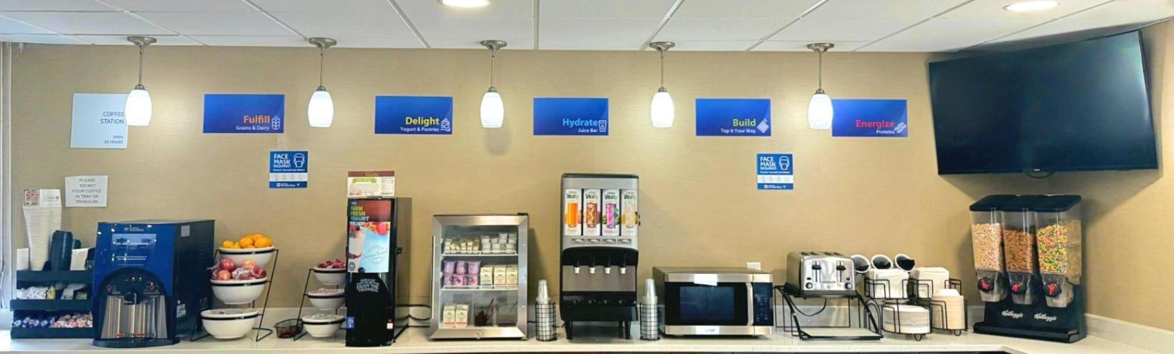 Un área de comida autoservicio con diferentes dispensadores de bebidas, un microondas y un refrigerador.