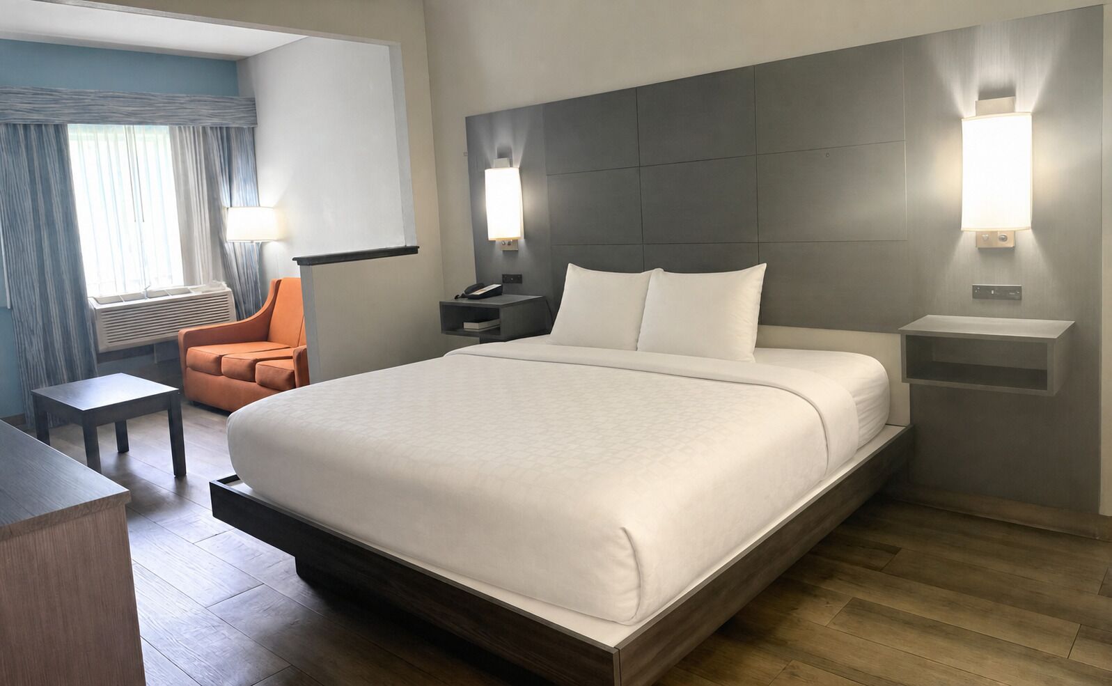 A hotel room with a large bed, bedside tables, a desk, an orange chair, and a view of the outdoors.