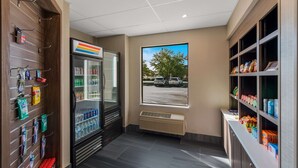 A clean, modern restroom with a refrigerated beverage display, a window with a parking lot view, and shelves stocked with snacks and toiletries. - Comfort Inn & Suites Tipp City Dayton North I-75 (Tipp City)