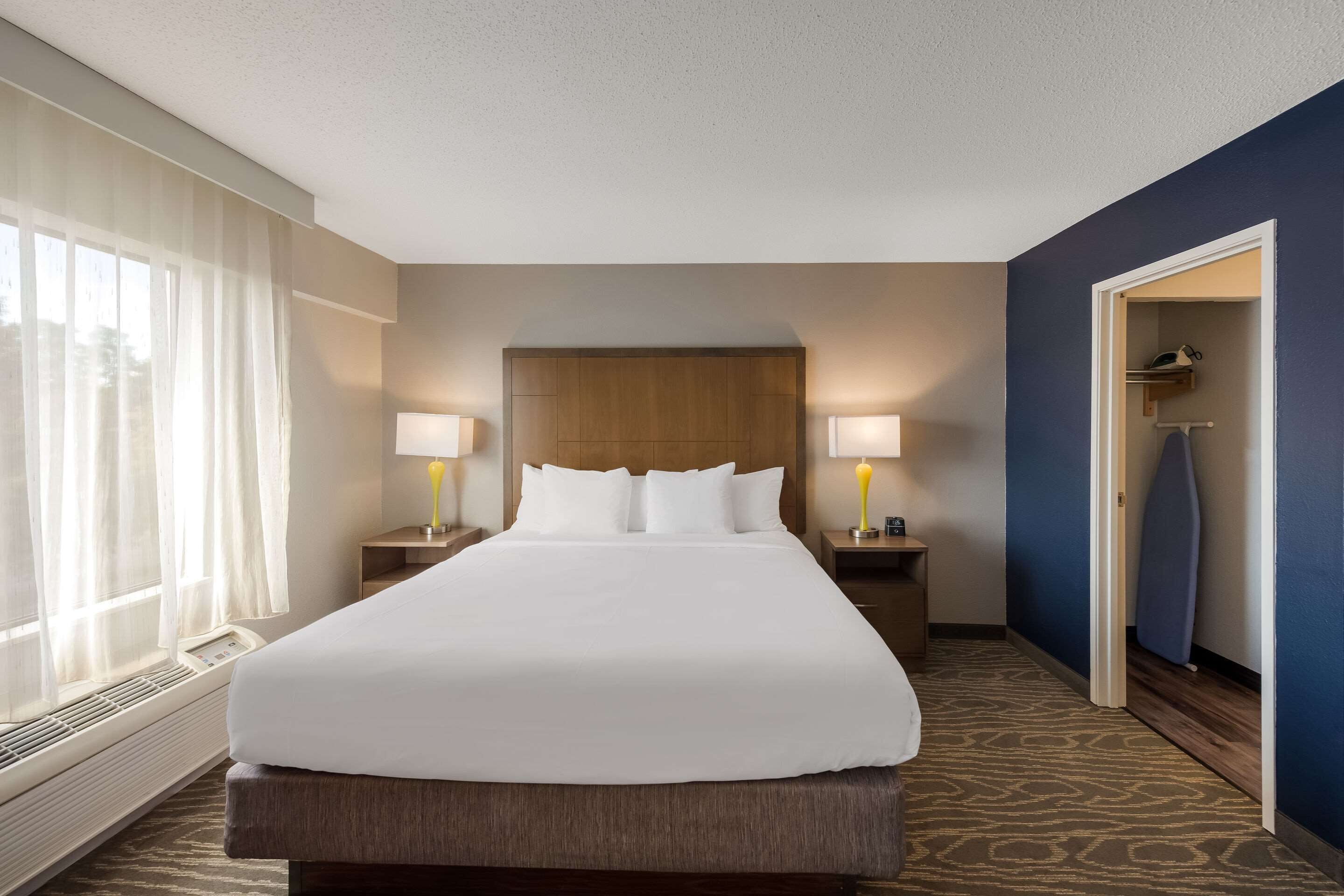 A hotel room with a large bed, two bedside lamps, a window with sheer curtains, a wooden headboard, and a blue accent wall.