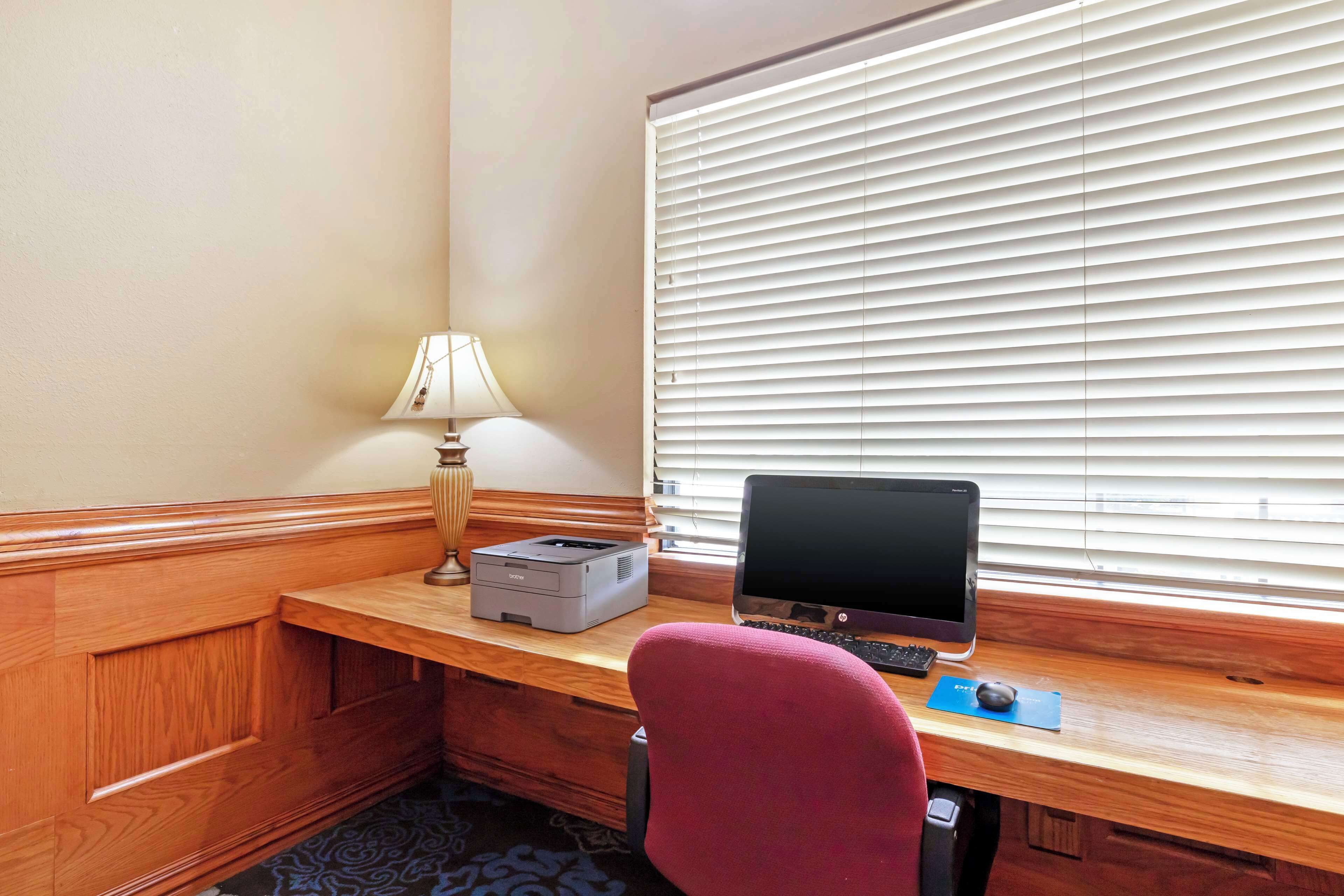 A hotel room with a wooden desk, a computer, a printer, a lamp, and a window with blinds.