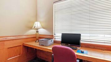 A hotel room with a wooden desk, a computer, a printer, a lamp, and a window with blinds.