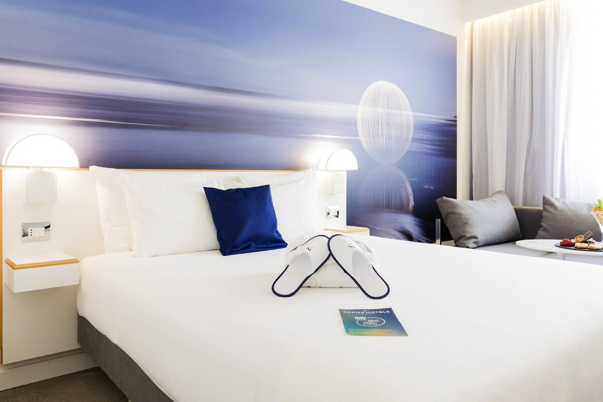 A hotel room with a large bed, a nightstand, a wall-mounted lamp, a pillow, a blanket, a pair of slippers, and a brochure on the bed.
