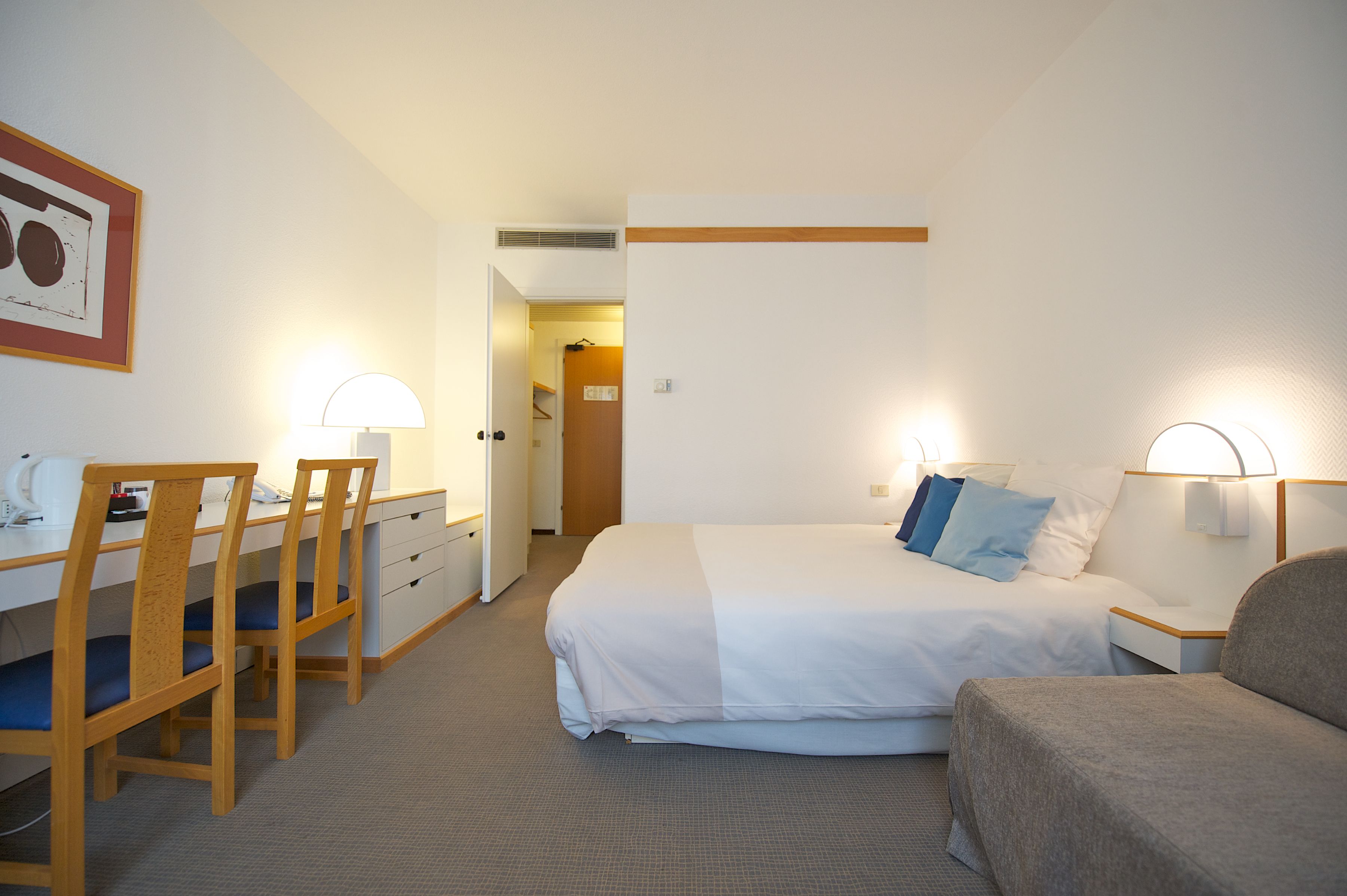 A hotel room with a bed, a desk with chairs, a lamp, and a framed picture on the wall.