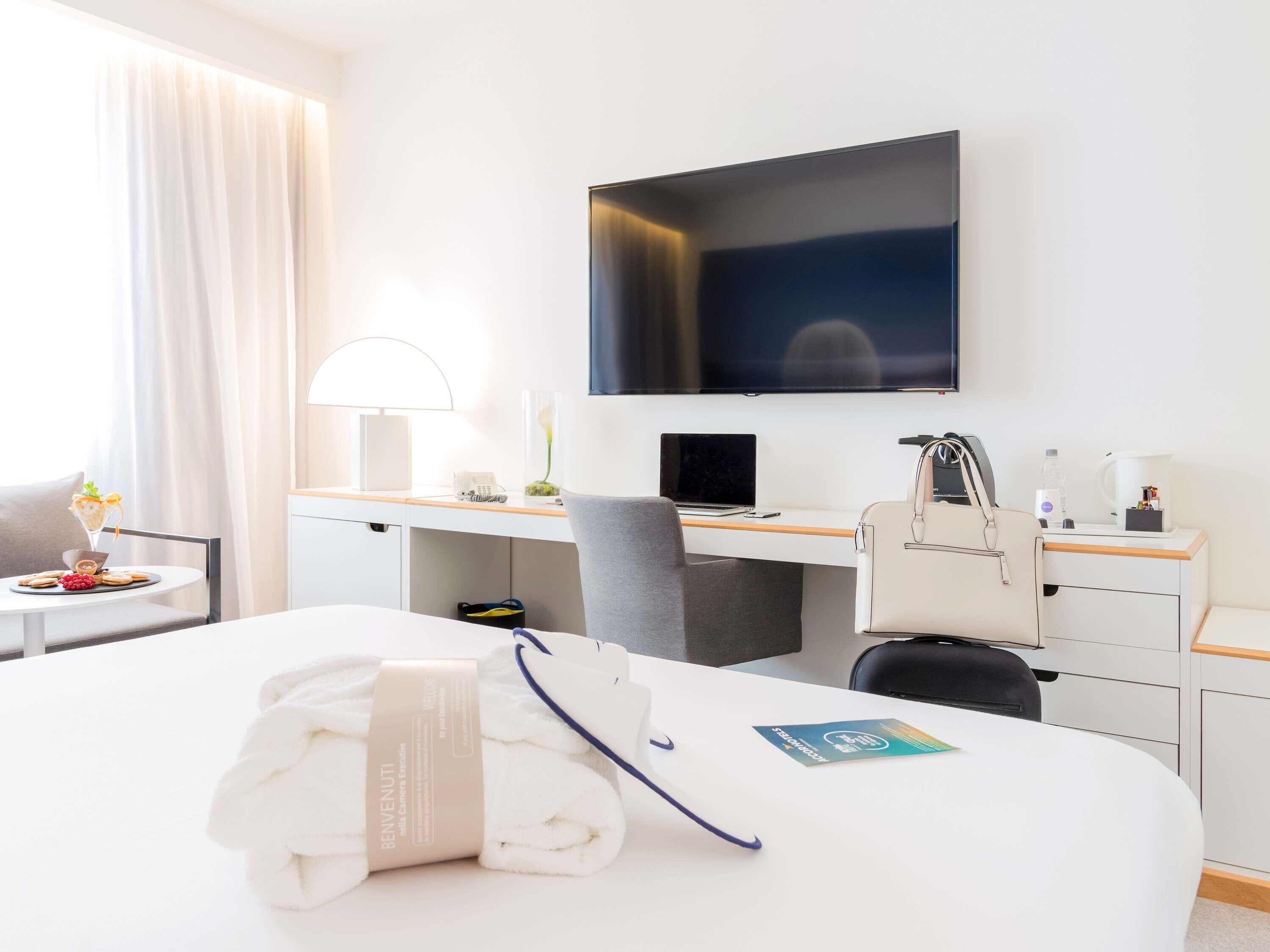 A hotel room with a bed, desk, chair, TV, and a small table with a drink and fruit.
