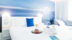 A hotel room with a large bed, white bedding, a blue pillow, a nightstand with a lamp, and a wall mural. - Novotel Firenze Nord Aeroporto (Sesto Fiorentino)