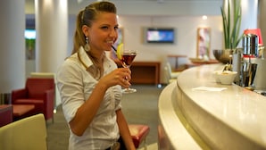 A person seated at a bar with a drink, a television in the background, and a red chair visible. - Novotel Firenze Nord Aeroporto (Sesto Fiorentino)
