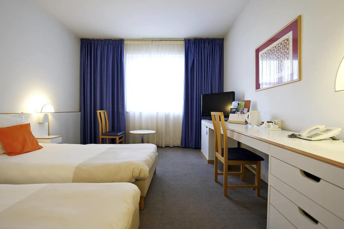 A hotel room with two beds, a desk with a computer, a chair, a telephone, and a window with curtains.
