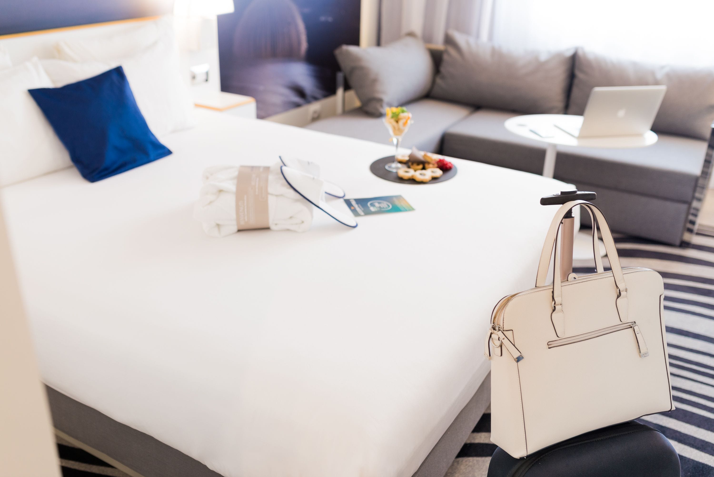 A hotel room with a bed, a laptop on a small table, a handbag, and a tray with food and a drink.
