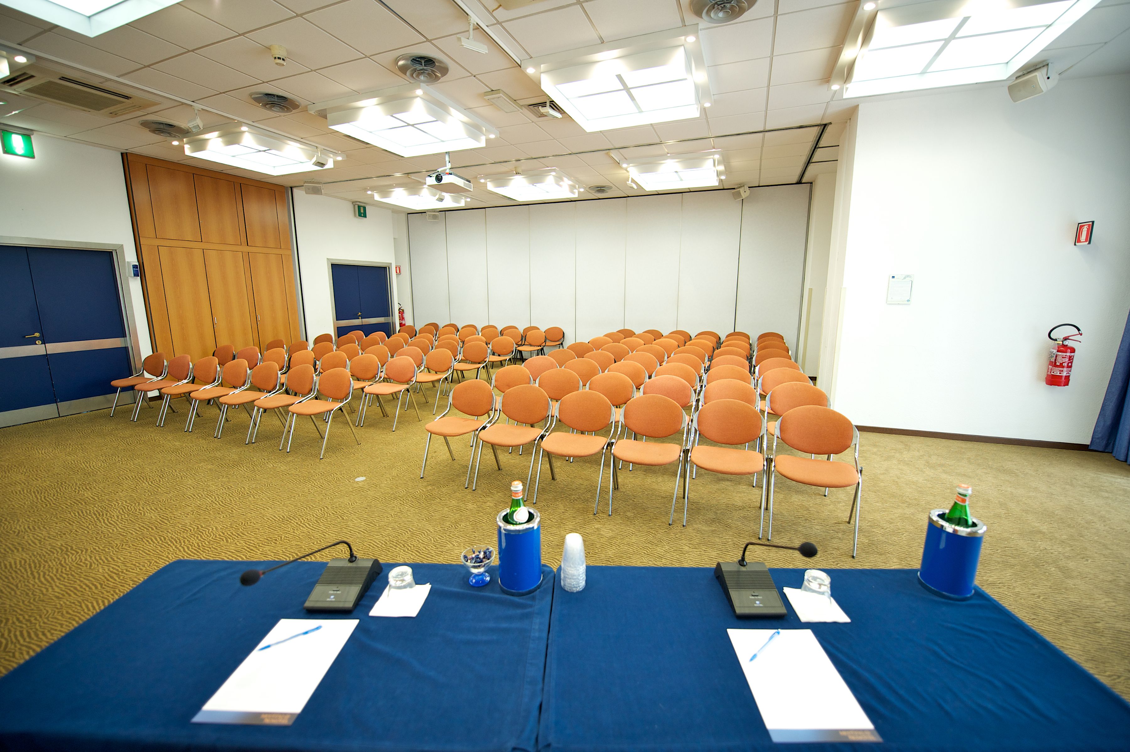 Ein Konferenzraum mit Reihen orangener Stühle, einem blauen Tisch mit Mikrofonen und Papieren sowie einem Feuerlöscher an der Wand.