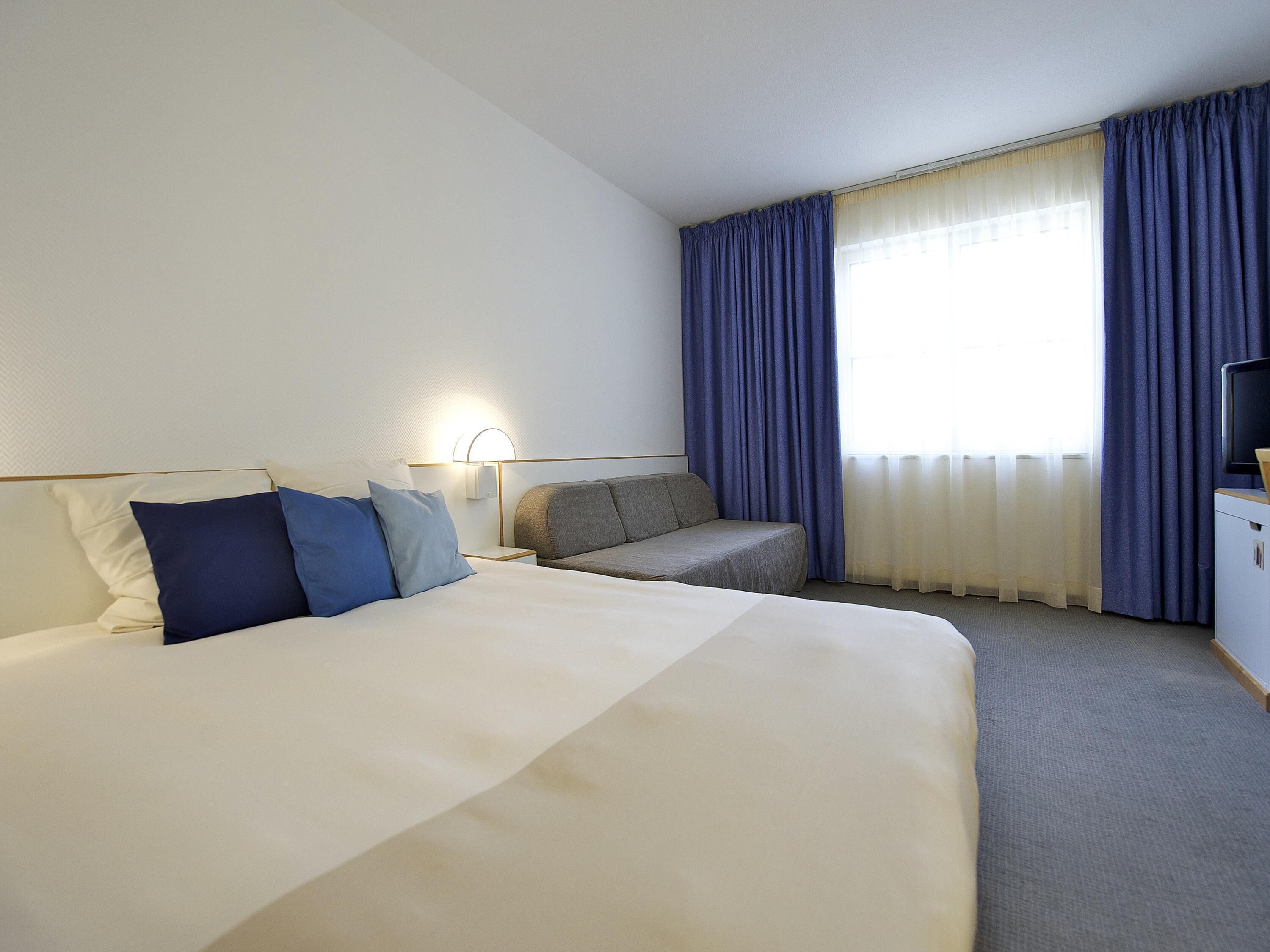 Quarto de hotel com uma cama grande, um sofá, uma escrivaninha e uma televisão.