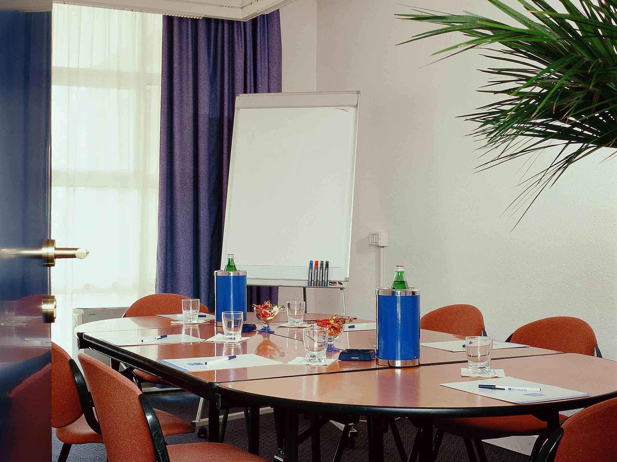 Uma sala de conferência com quadro branco, mesa com cadeiras, planta e janela com cortinas.