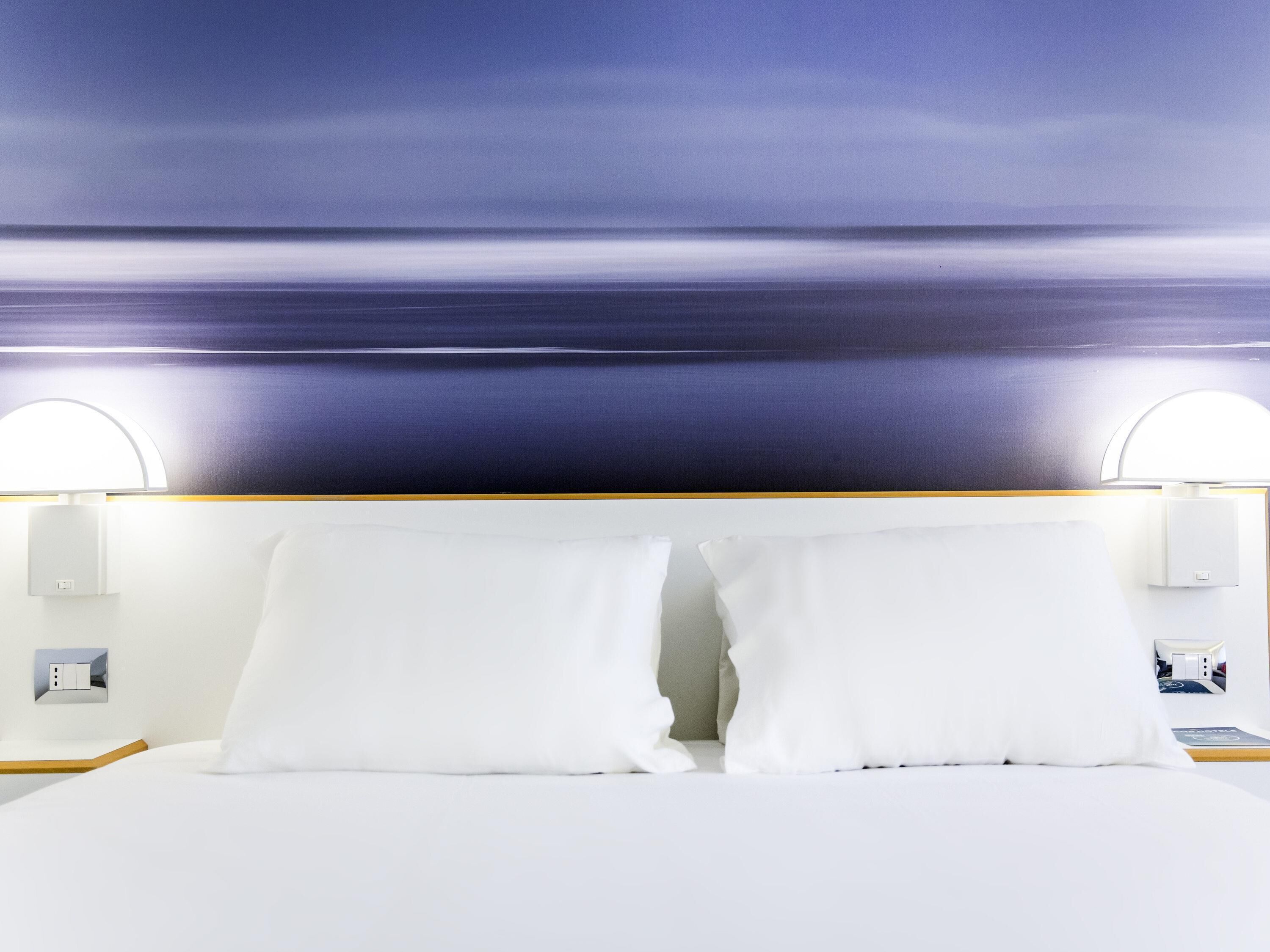 A hotel room with a bed, white pillows, a nightstand, a wall-mounted lamp, and a wall-mounted power outlet.