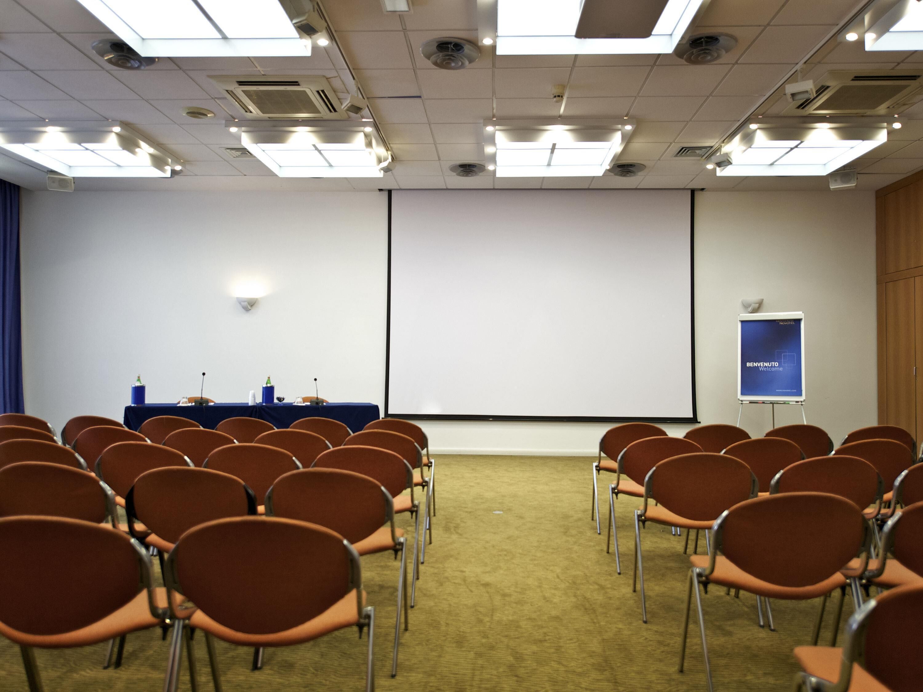 A conference room with rows of chairs, a projector screen, a presentation screen, and a whiteboard.