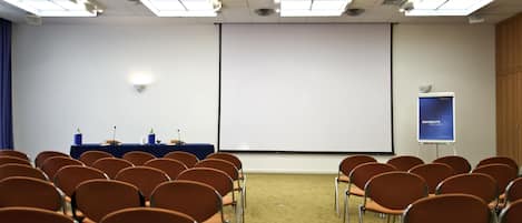 A conference room with rows of chairs, a projector screen, a presentation screen, and a whiteboard.