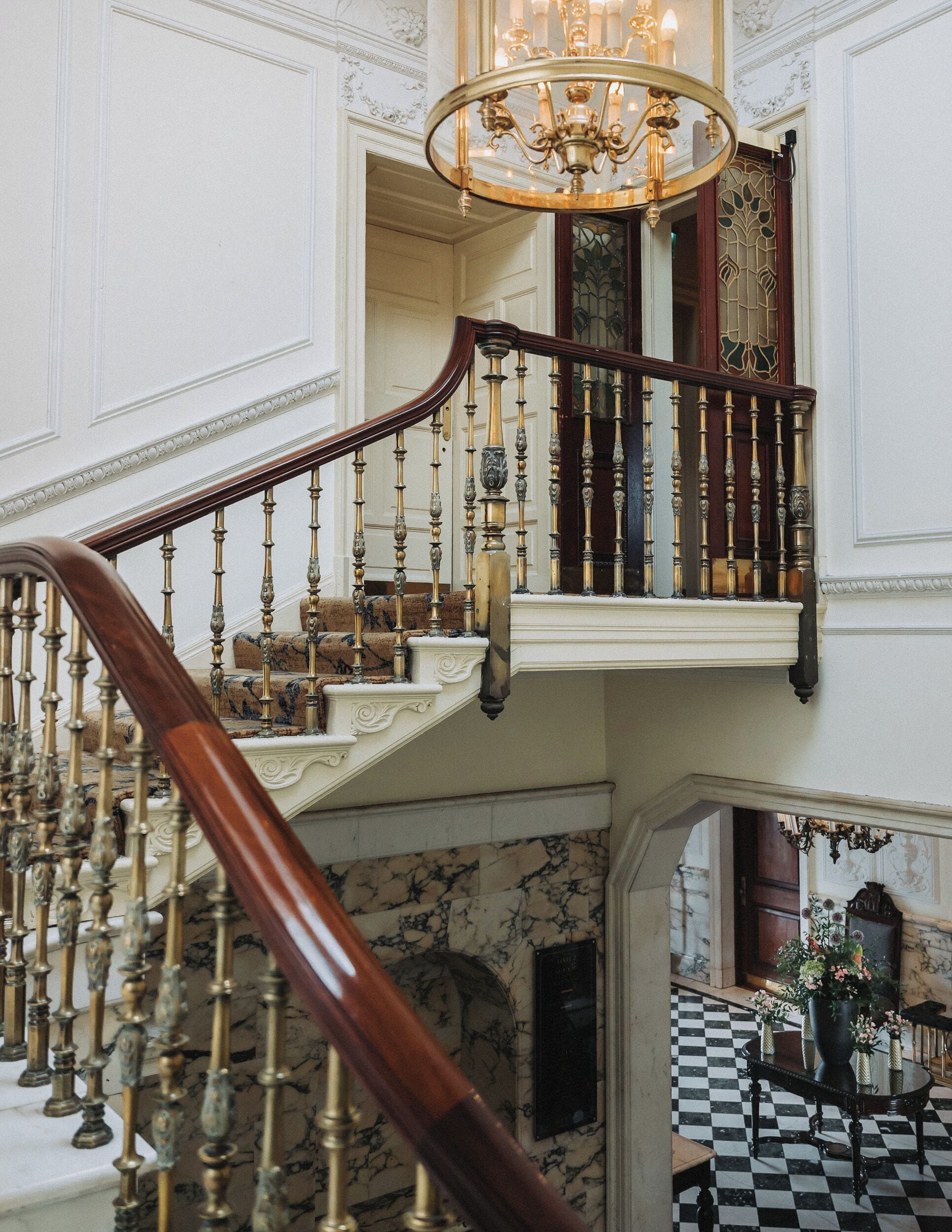 A grand staircase with ornate balustrades and a chandelier, leading to a balcony with a stained glass window.