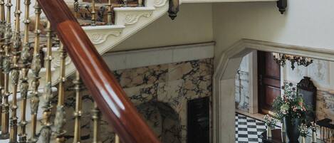 A grand staircase with ornate balustrades and a chandelier, leading to a balcony with a stained glass window.