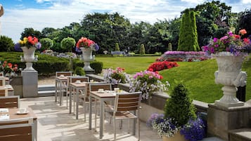 Un área de estar al aire libre con mesas y sillas, rodeada de jardineras con flores y un césped cuidado.