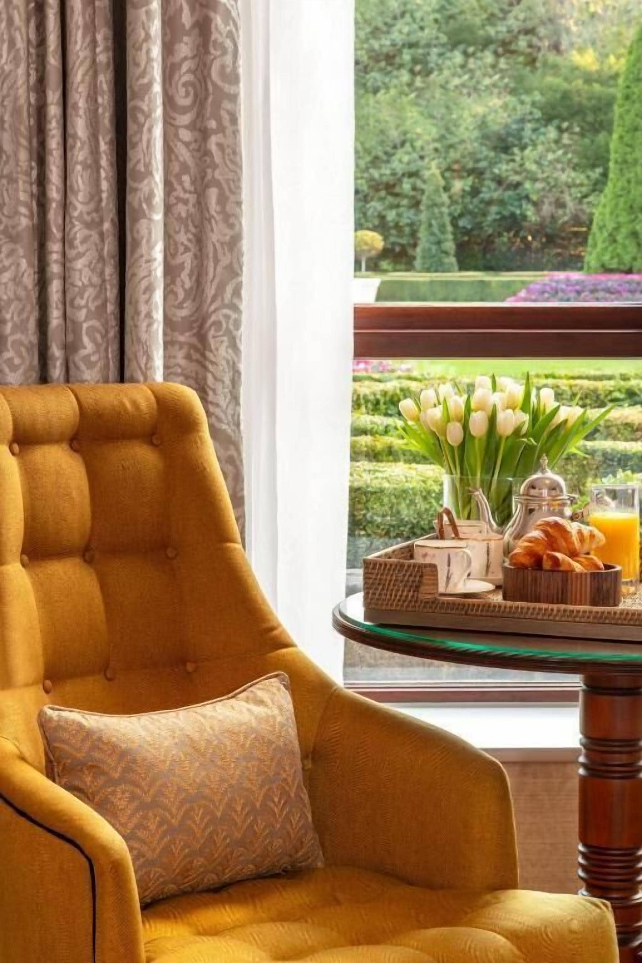 A room with a tufted armchair, a window with a view of a manicured garden, and a breakfast tray on the windowsill.