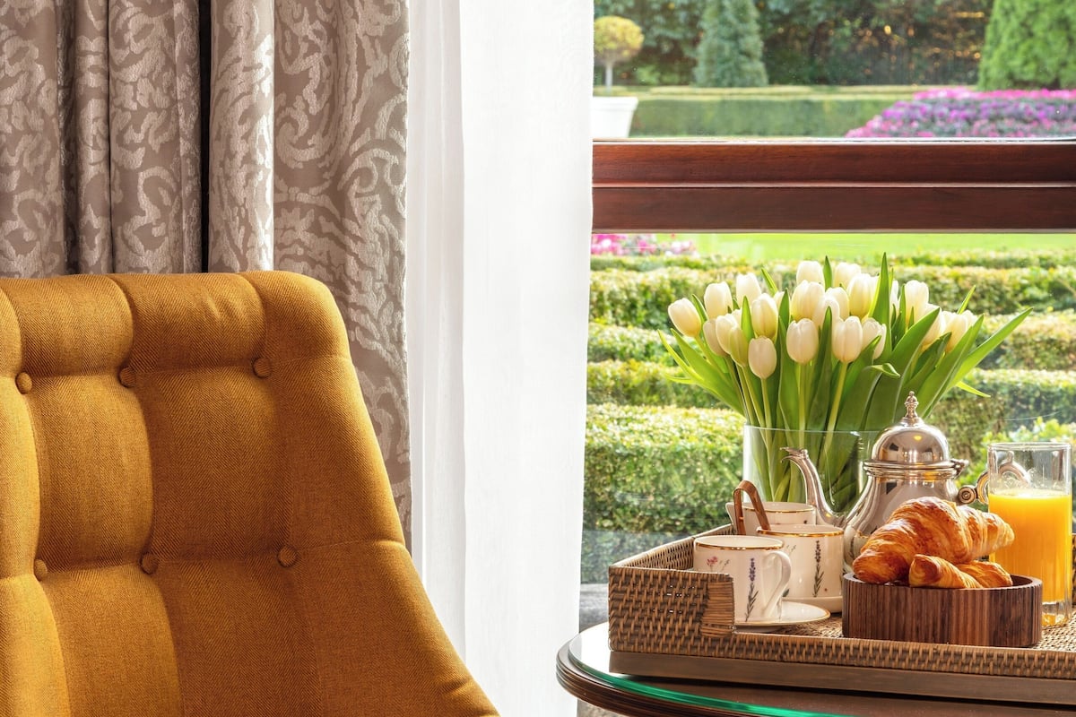 A mustard-colored armchair with a patterned cushion, a glass table with breakfast items, and a view of a well-manicured garden through a window.