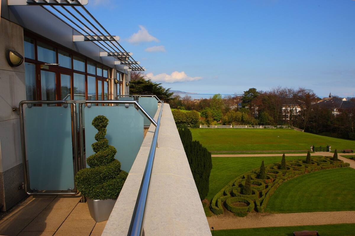 A modern building with a glass facade, a balcony with metal railings, and a well-maintained garden with hedges and a lawn.