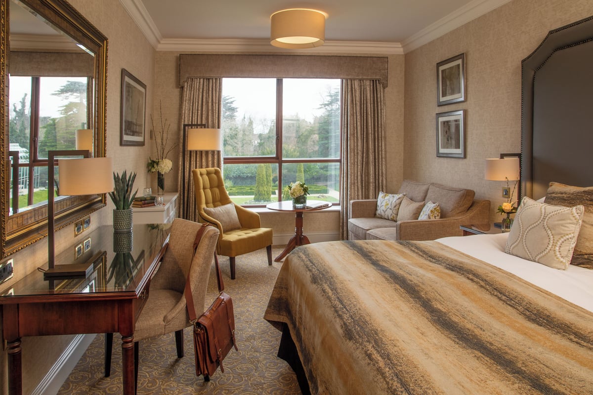 A hotel room with a large bed, a sofa, a chair, a desk, and a large window offering a view of the outdoors.