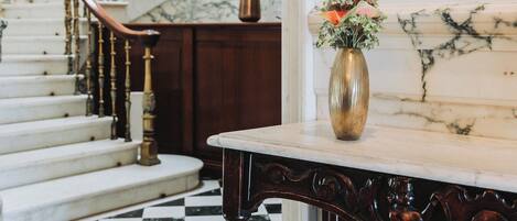 A grand staircase with a checkered marble floor, a wooden railing, and a vase with flowers on a marble table.