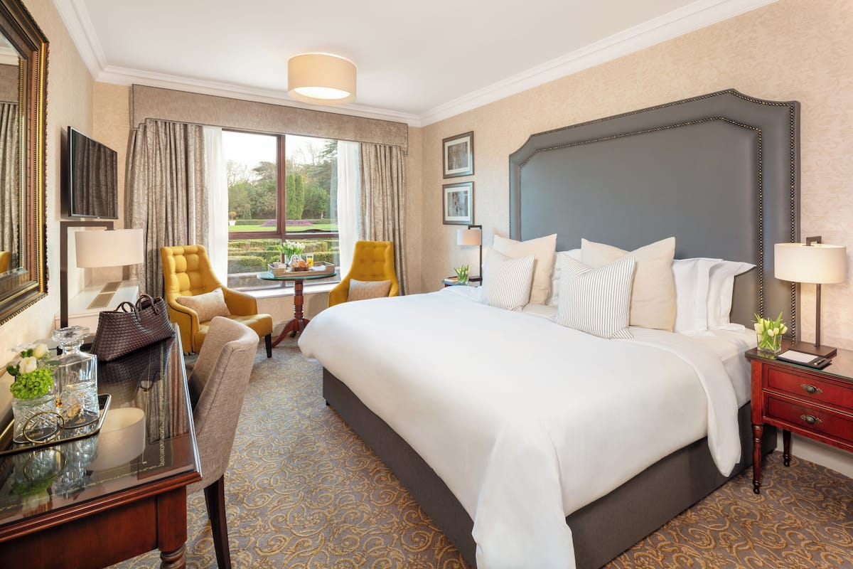 A hotel room with a large bed, a sitting area with a chair, a desk with a lamp, and a view of a garden through the window.