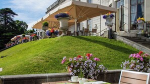 Un patio exterior con una mesa lista para comer, rodeado de sillas, y vistas a un césped cuidado y un edificio clásico con escalones que conducen a la entrada.