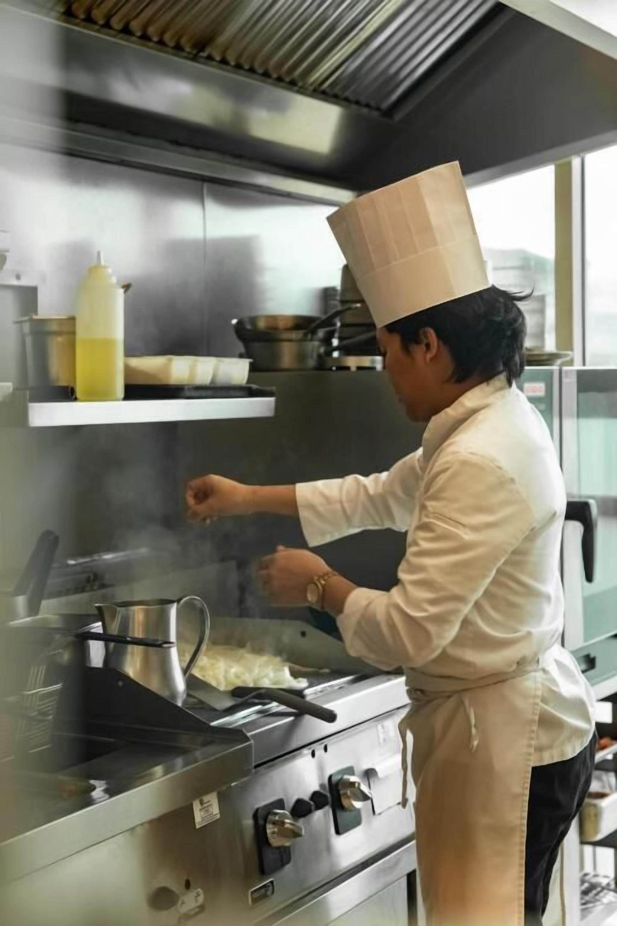 Un chef con uniforme y gorro blanco cocina en una cocina de acero inoxidable con una ventana.