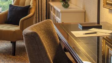 Habitación de hotel con un ventanal grande, un sillón cómodo, un escritorio con lámpara y una mesita con un jarrón de flores.