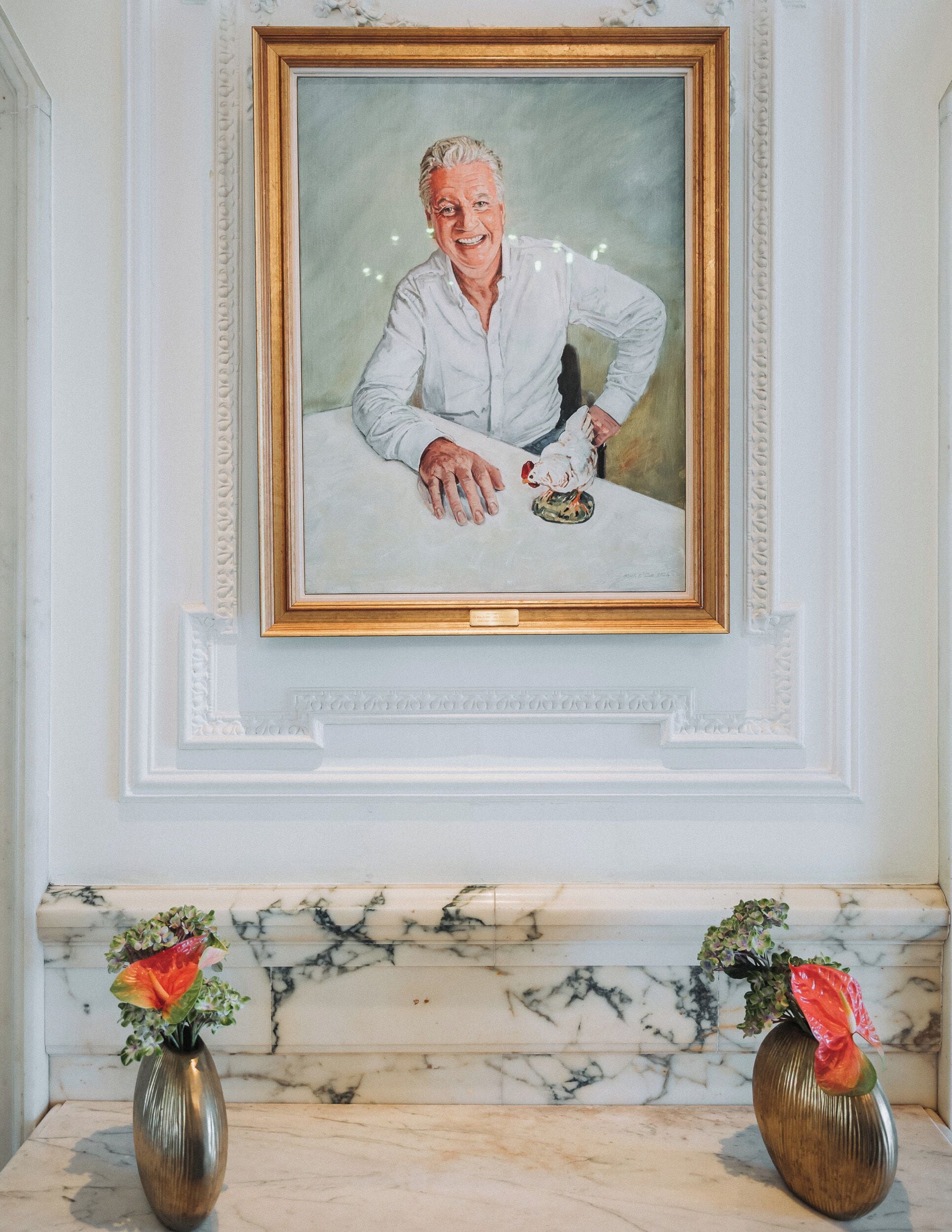 A framed portrait of a person seated at a table with a small bird on it, flanked by two vases with flowers on a marble mantelpiece.