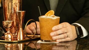 A person in a black suit holding a cocktail glass with an orange garnish, seated at a bar with copper-colored glasses and a watch on their wrist.