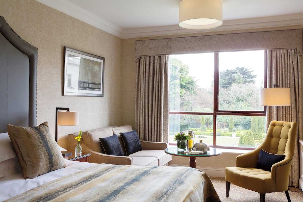 A hotel room with a bed, two armchairs, a sofa, a small table with a vase of flowers, and a window with curtains.
