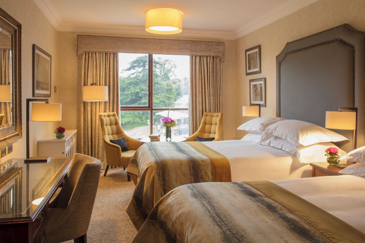 A hotel room with two beds, a sitting area with a chair, a desk, and a large window with curtains.