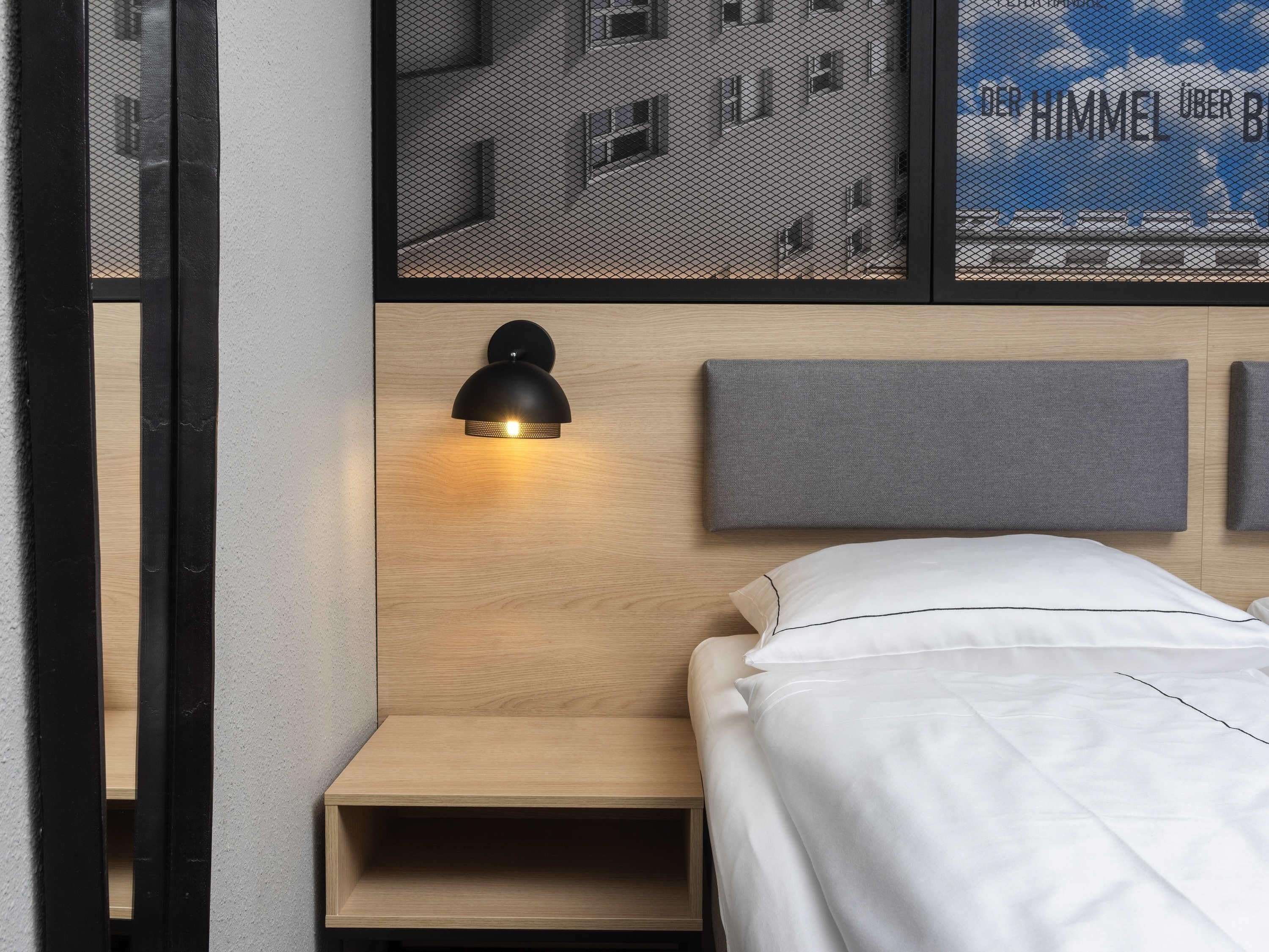 A modern hotel room with a wooden headboard, a bedside table, a bed with white bedding, and a wall-mounted lamp.