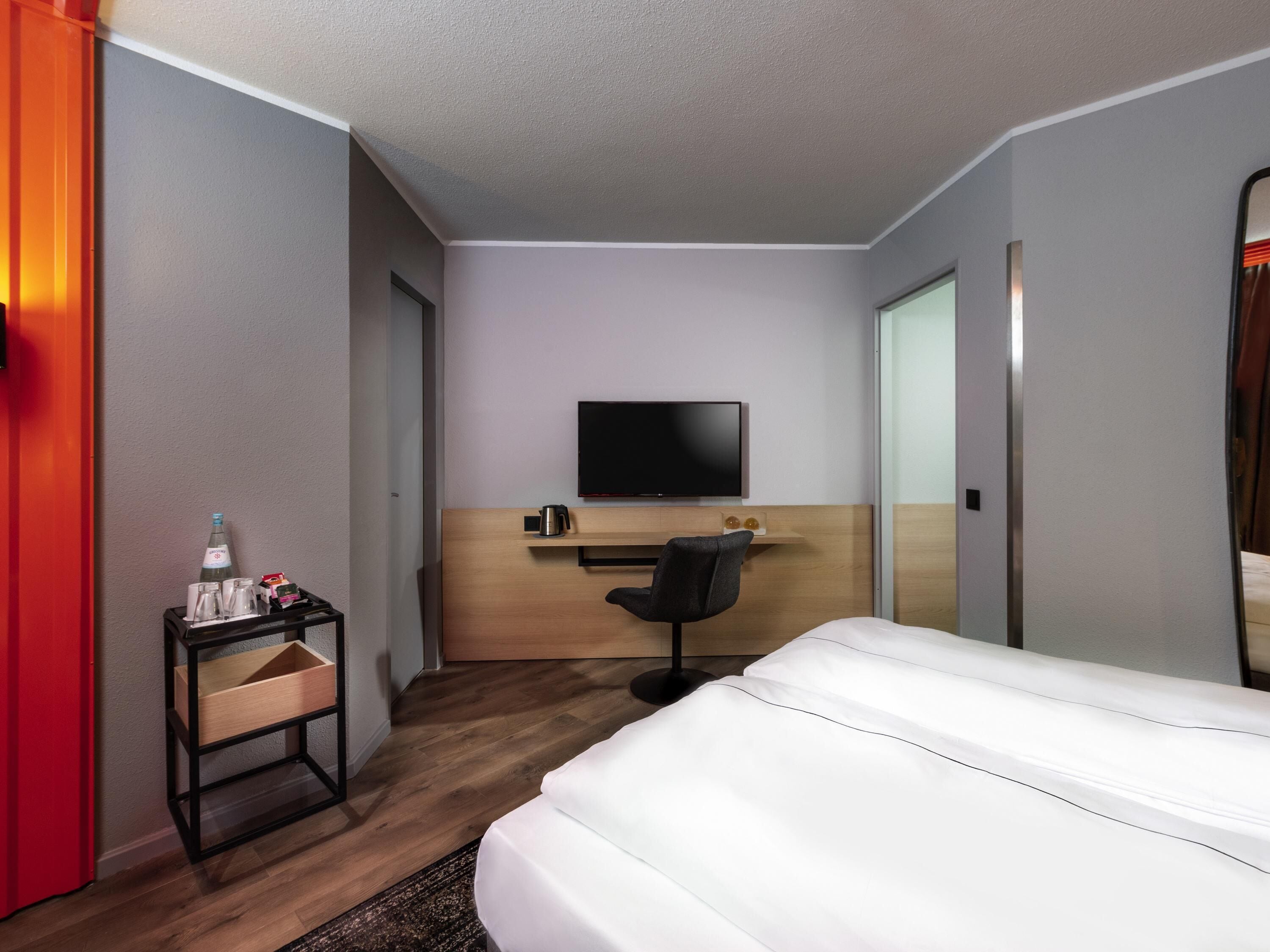 Quarto de hotel com uma cama, uma escrivaninha, uma cadeira, uma televisão e uma pequena mesa com uma garrafa e copos.