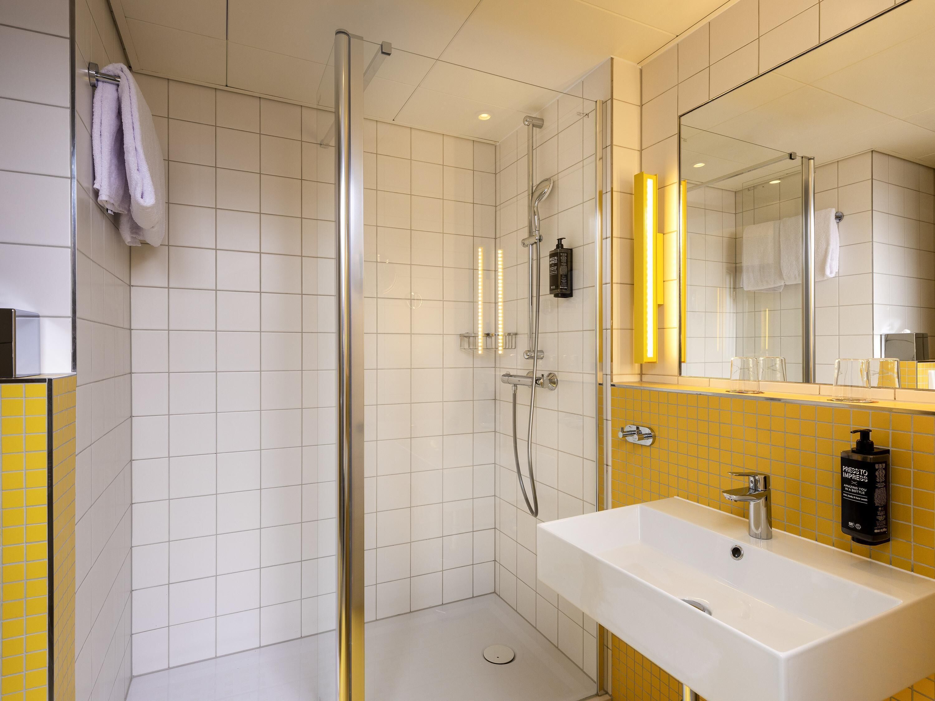 Un baño moderno con ducha envuelta en vidrio, un lavamanos blanco y una pared de acento amarilla, y un espejo con una tira de luz amarilla.