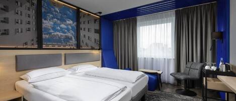 Habitación de hotel moderna con una cama grande, un escritorio y una silla. Se ve una ventana con cortinas y vistas a edificios.