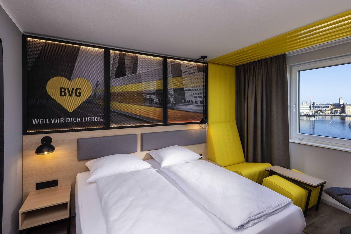 Ein Hotelzimmer mit Bett, Schreibtisch, Stuhl, Fenster mit Blick auf eine Stadtansicht und einer Wand mit Stadtbild und Text.