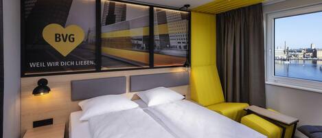 Habitación de hotel con cama, escritorio, silla, ventana con vista a un paisaje urbano y pared con un paisaje urbano y texto.