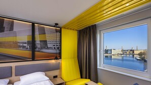A hotel room with a bed, a yellow armchair, a small table, and a view of a river and buildings. - ibis Styles Berlin Treptow (Berlin)