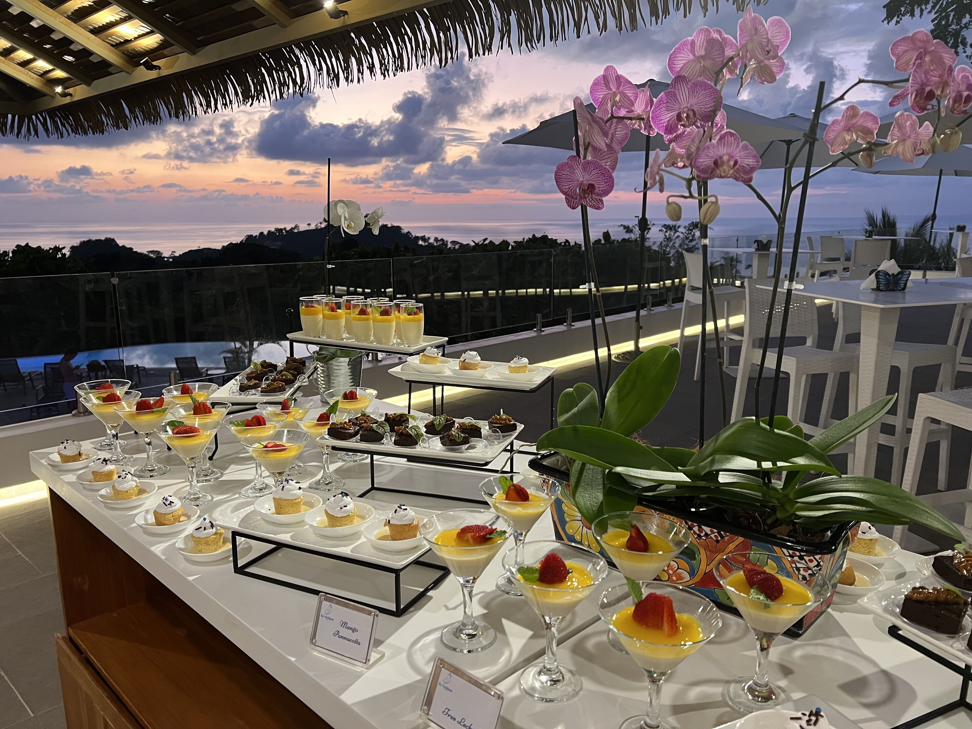 Een dessertbuffet met diverse gepresenteerde desserts en drankjes, met op de achtergrond een zonsondergang en tropische bloemen.