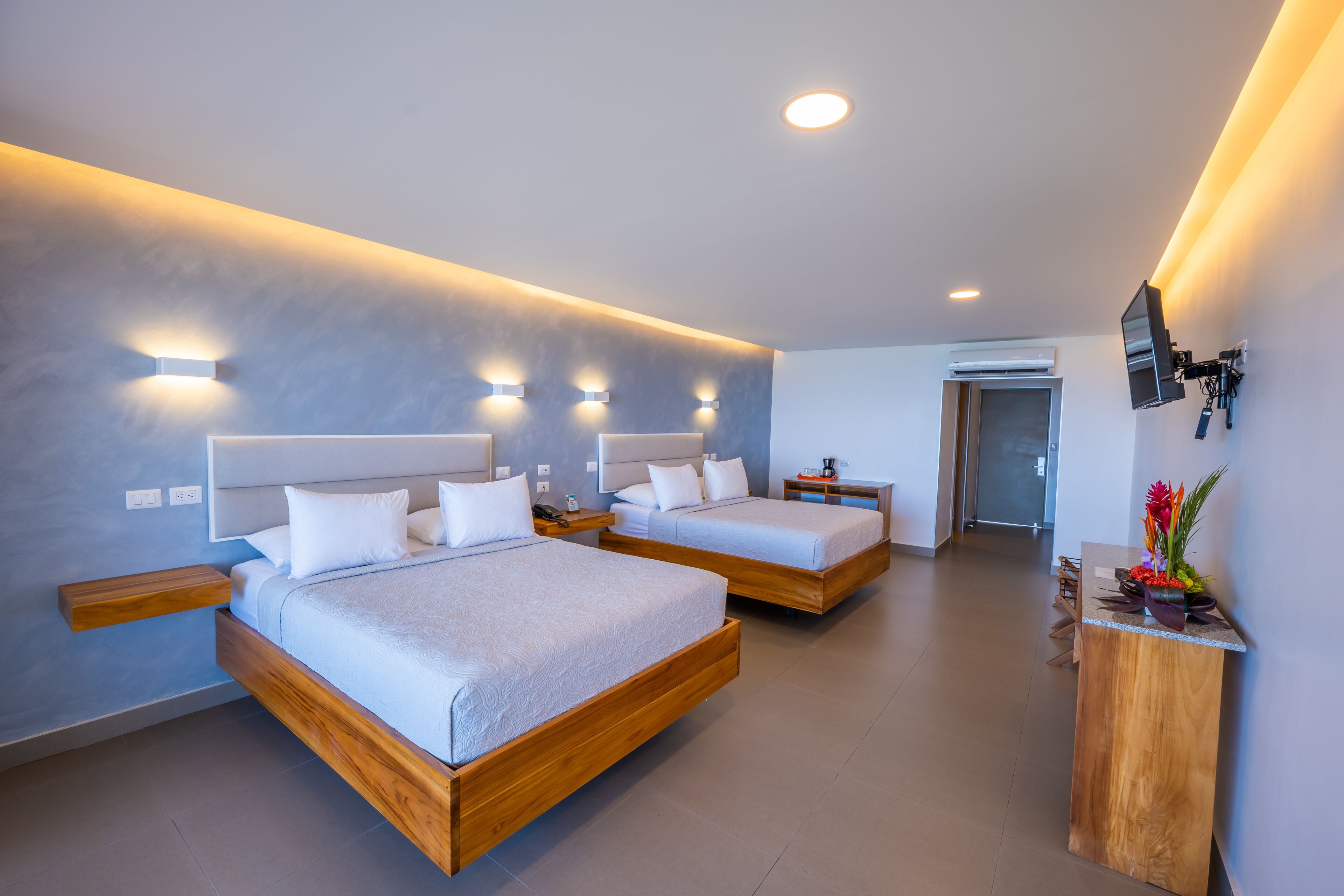 A modern hotel room with two wooden beds, a wooden nightstand, a flat-screen TV mounted on the wall, and a small wooden table with a vase of flowers.