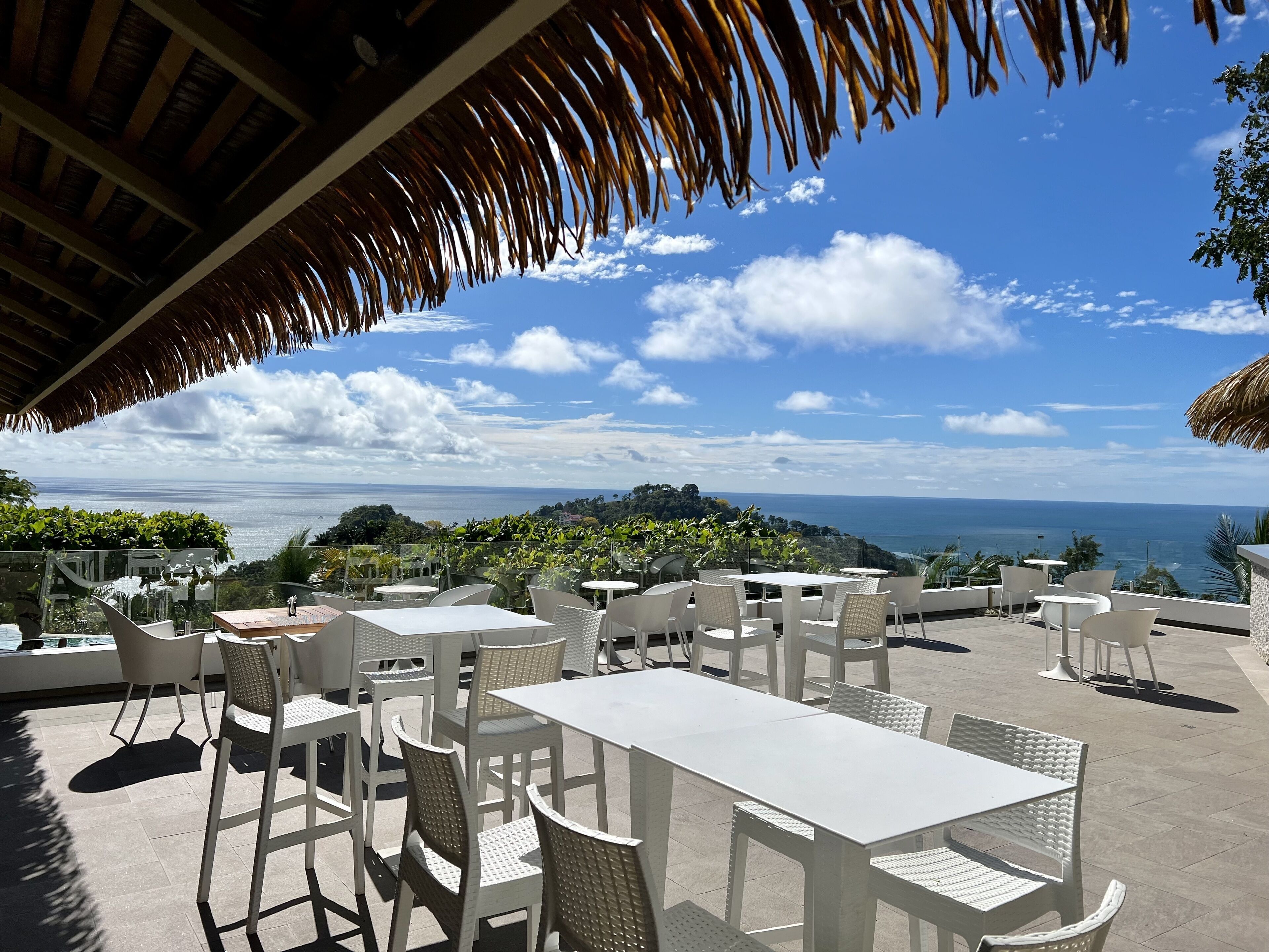 Un espace extérieur aménagé avec des tables et des chaises blanches, offrant une vue panoramique sur la mer et les collines.