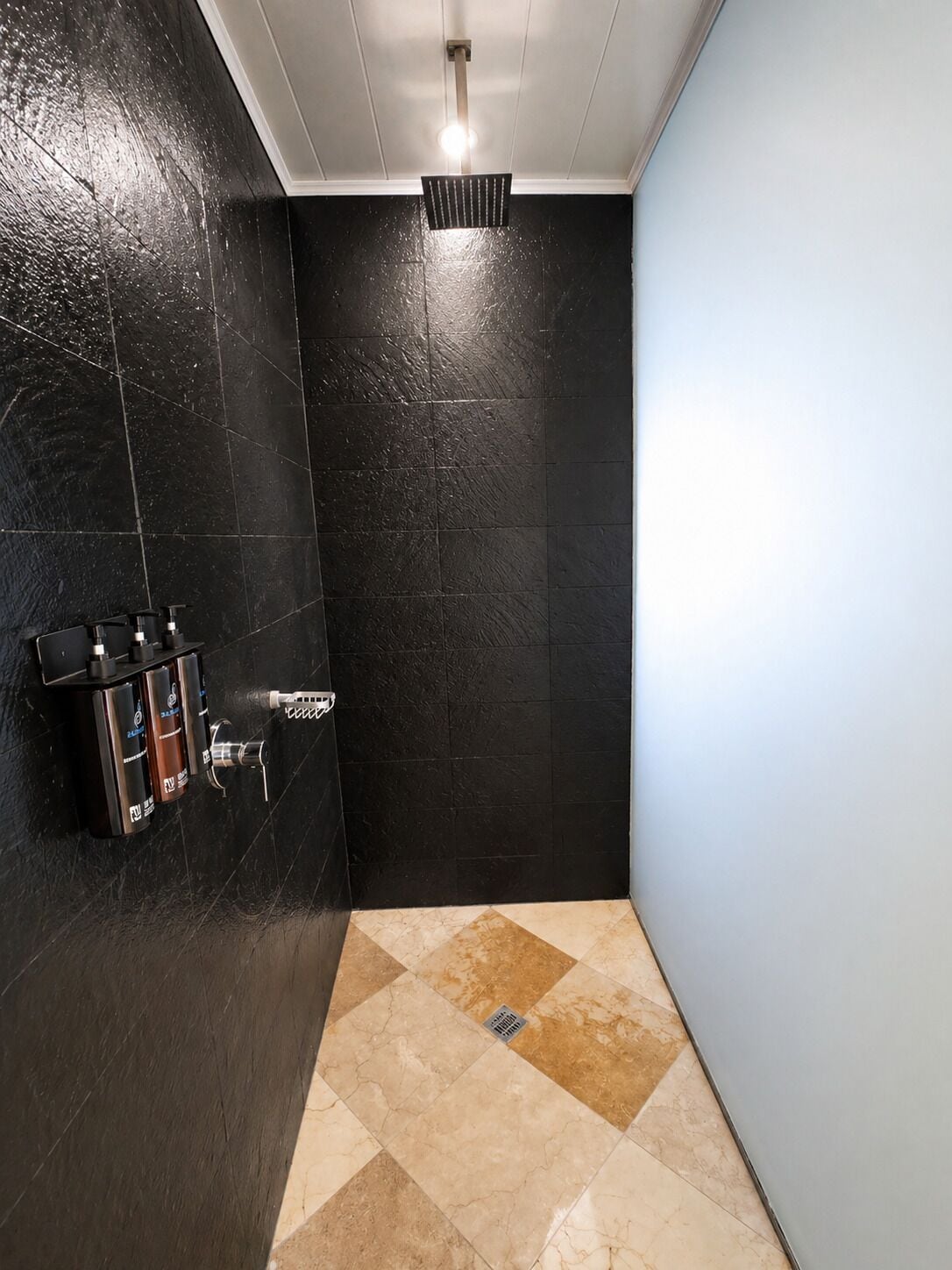 A bathroom with black tiled walls, a showerhead, and a hand soap dispenser.
