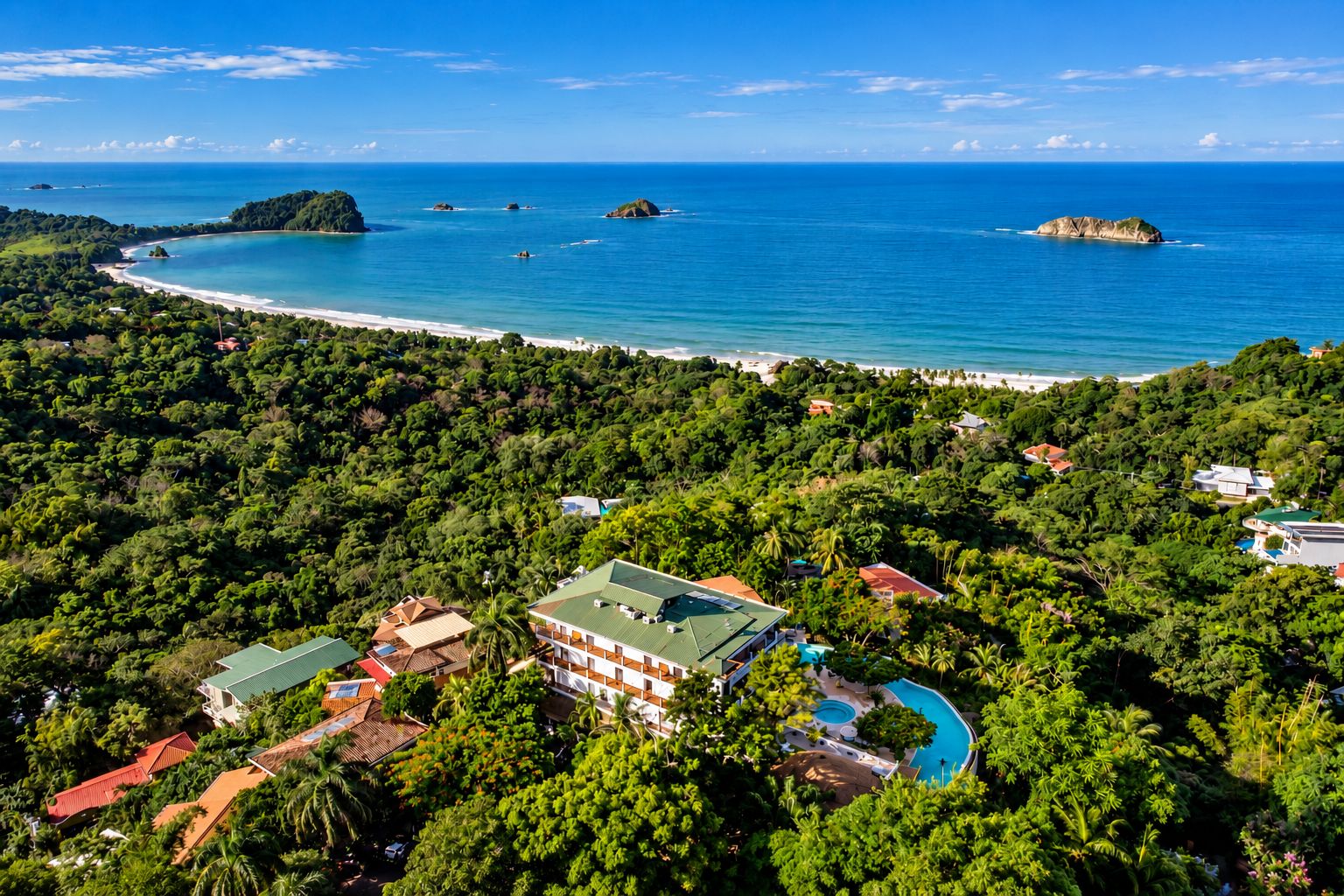 An aerial view of a coastal resort with a swimming pool, surrounded by dense tropical vegetation and a beach.