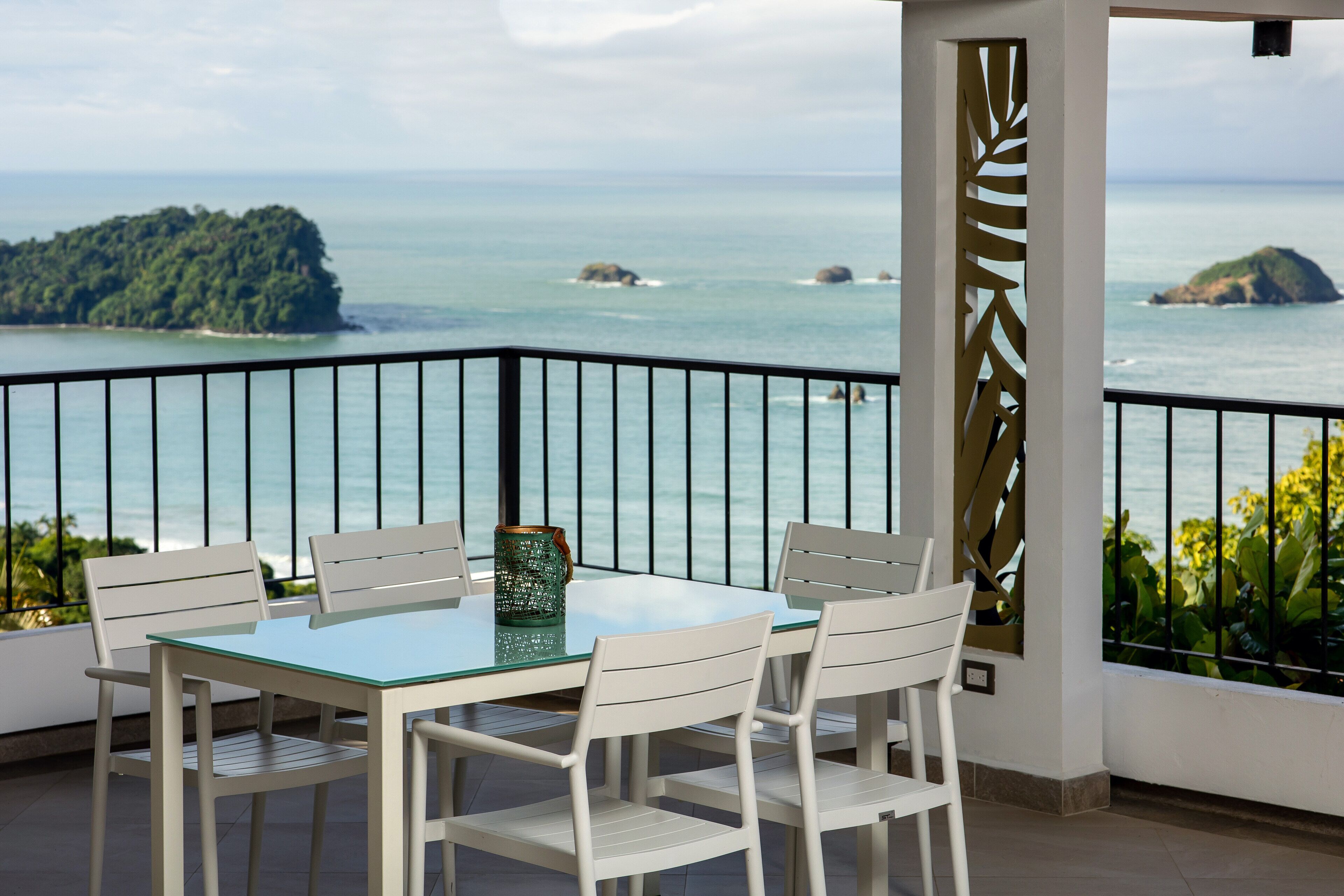 A balcony with a glass table and white chairs, overlooking a coastal view with rocky islands and the sea.